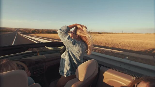 View from back seat as young women enjoy lifestyle during ride in convertible, women in sunglasses raise their hands, have fun and fool around. Ride in convertible on highway, sunset