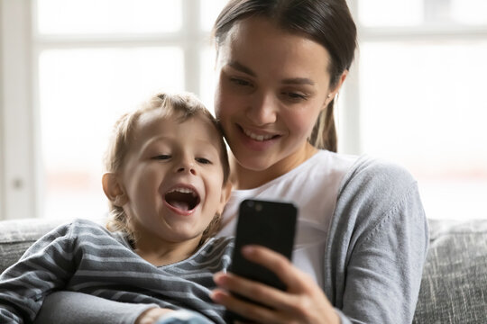 Happy mum and excited cute son kid speaking and smiling at smartphone screen, resting on sofa at home. Mother hugging child, using mobile phone for video call, taking selfie, shopping online