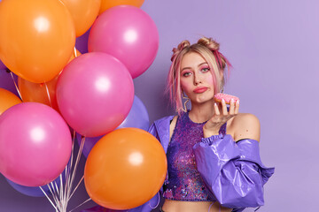 Fototapeta na wymiar Serious attractive young European woman looks bored at camera dressed in fashionable purple jacket and cropped top holds tasty donut being on party poses with bunch of colorful helium balloons