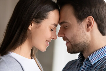Happy millennial couple in love touching with foreheads, smiling with closed eyes. Close up faces of affectionate man and woman enjoying romantic intimate moment, hugging, expressing tender.