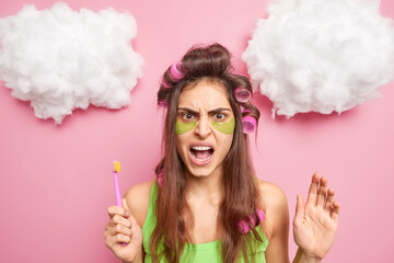 Displeased angry dark haired woman applies hair curlers collagen patches under eyes to reduce...