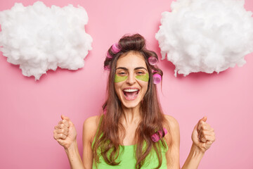 Positive woman clenches fists celebrates something applies hair rollers green beauty patches under eyes wants to have great haircut tomorrow treats herself isolated on pink background. Skin treatment