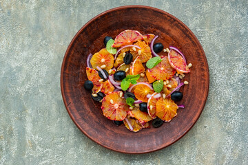A healthy salad of sliced bloody oranges, purple onions, olives and pine nuts with olive oil dressing on a brown plate against an gray concrete background. Vegan recipes.