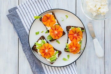 A healthy breakfast or snack, open sandwiches on rye bread with cottage cheese or cream cheese, slices of bloody oranges and herbs on a white plate