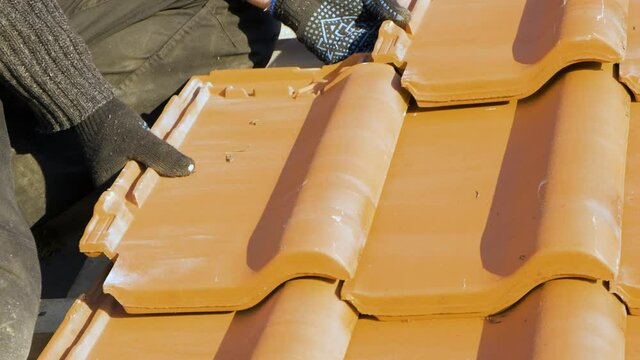 Closeup of worker hands installing yellow ceramic roofing tiles mounted on wooden boards covering residential building roof under construction.