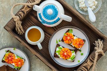 Fototapeta na wymiar A healthy breakfast or snack, open sandwiches on rye bread with cottage cheese or cream cheese, slices of bloody oranges and herbs on two plates on a gray concrete background.