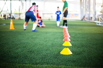 Cone markers is soccer training equipment on green artificial turf with blurry kid players training background. Material for training class of football academy.