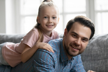 Portrait of cute girl lying on daddy back, looking at camera and smiling. Happy father lying on sofa, piggybacking daughter, having fun with kid. Best dad enjoying leisure with child at home. Closeup