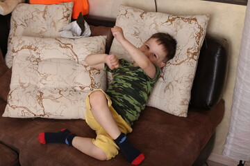 baby boy sitting on a brown sofa in yellow shorts, green T-shirt, playing around, covering his face with his hands