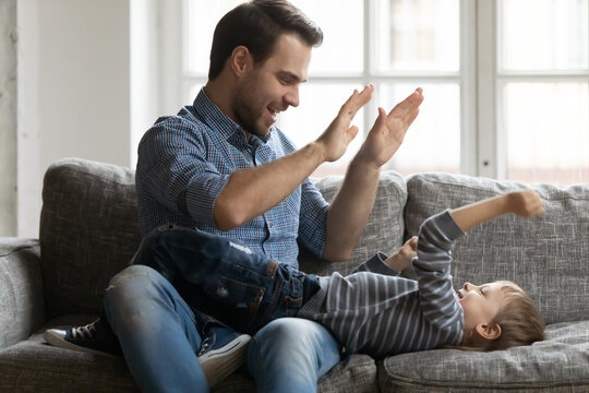 Happy Father Giving High Five To Cute Preschooler Son With Both Hands. Best Dad Cuddling Boy Kid On Sofa In Living Room, Relaxing And Enjoying Leisure Time With Child At Home. Family Playtime Concept