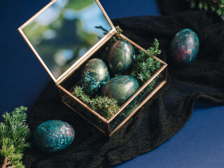 Mother of pearl Easter eggs lie in a glass box and a blue background. There are twigs of juniper...
