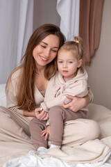 Fototapeta na wymiar Portrait of smiling young mommy embracing little daughter. Mother with child sitting on bed and hugging. Close up