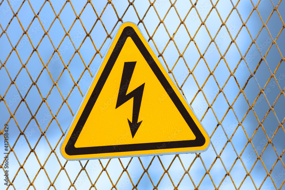 Poster warning sign, yellow triangle with lightning on a fence made of metal mesh