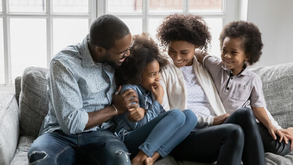 Happy millennial black married couple and two kids enjoying leisure time in living room together. African American parents and children relaxing and hugging on couch at home. Family bonds concept