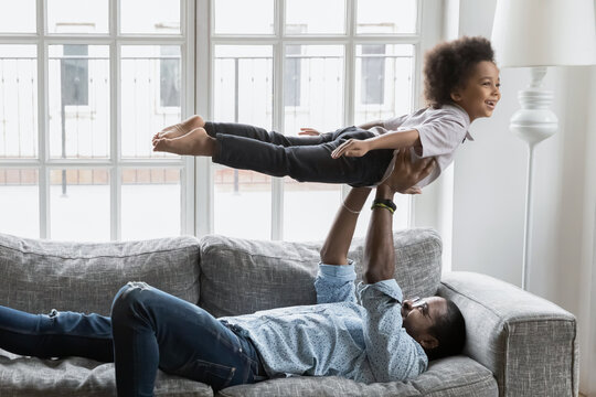 Happy father and little son doing together funny exercises at home. Dad resting on couch, lifting up, holding boy in air, play active game. Excited kid making airplane or boat. Family activity concept
