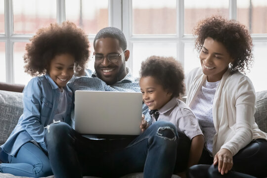 Happy African American couple and two kids relaxing on couch at home, watching funny movie together, laughing. Black family using laptop pc for internet call, distance talk, video chat
