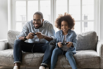 Excited girl and her dad play video game together, sitting on couch, using controllers. Black...