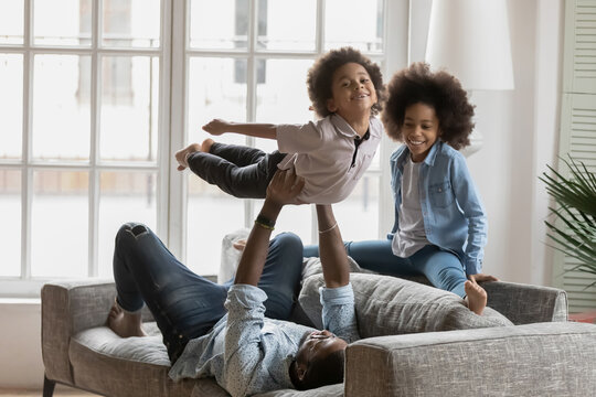 African American father playing funny active sport games with kids at home. Happy children and daddy having fun and doing exercises on couch. Dad lifting excited boy up in air. Sporty family activity