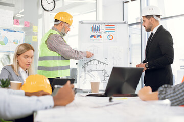 Male and female colleagues in suits and helmets analysing blueprints, graphs and charts of new project. People of different ages, and races. Concept of unity and cooperation.