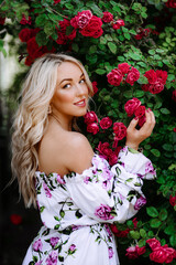 Naklejka premium Outdoor photo of romantic young woman in rose garden. girl with red lips in a dress with a print of roses. Young model on a background of a bush of roses. Stylish woman.