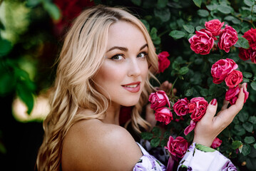 Outdoor photo of romantic young woman in rose garden. girl with red lips in a dress with a print of roses. Young model on a background of a bush of roses. Stylish woman.