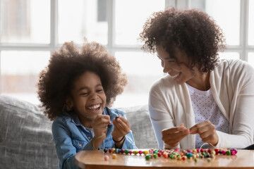 Happy excited mother and daughter kids sharing craft hobby, playing game, stringing colorful beads and laughing. Mommy and kid creating handmade decoration together, having fun at home. Candid moment