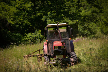 Obraz premium an old red tractor mowing the grass near the forest. use of agricultural machinery on the farm