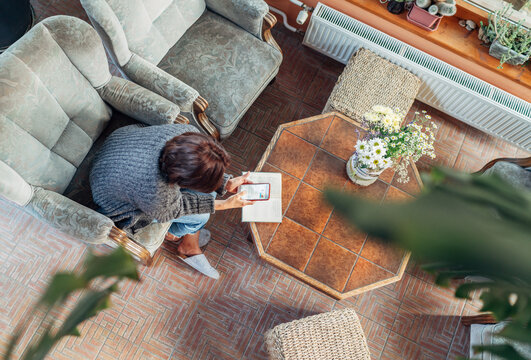 Top view of a female dressed cozy home clothes in comfortable armchair, browsing an internet using smartphone in house sunroom living room. Stay-at-home or business home office concept image
