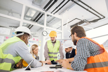 Fototapeta na wymiar Multiracial architect people in helmets sitting at table with blueprints and modern devices. Focused workers cooperating to create new building project.
