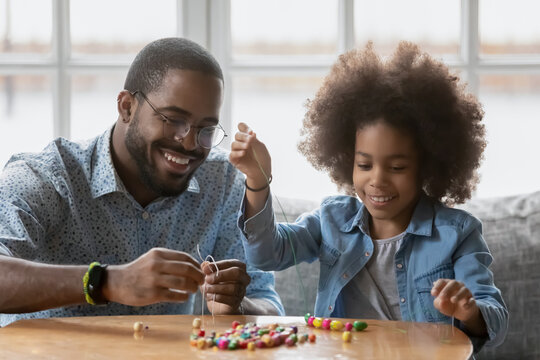 Happy Black father and daughter girl making decoration together, stringing colorful wooden beads, enjoying handmade activity. Daddy and kid improving craft skills, spending leisure time at home