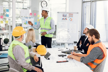 Group of international architects, designers and engineers sitting at office and working on common project. Afro-american man in helmet standing near flipchart and giving presentation to partners.