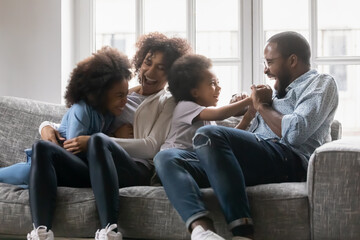Fototapeta premium Happy black married couple and kids having fun, tickling on couch in living room. Mom, dad and two kids enjoying leisure at home, sitting on sofa, hugging, cuddling, laughing. Family with children