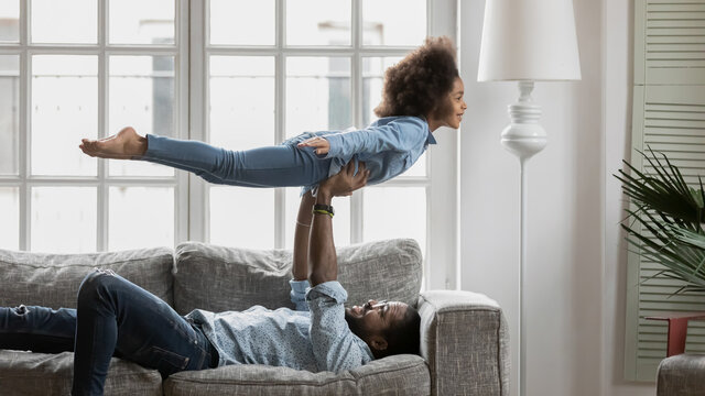 Happy strong father and little daughter enjoying funny activities in living room. Dad lying on sofa, lifting girl in air, kid playing flying plane. Family active games, home exercises concept