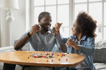Excited girl and happy dad stringing colorful wooden beads together. Young father helping kid to...