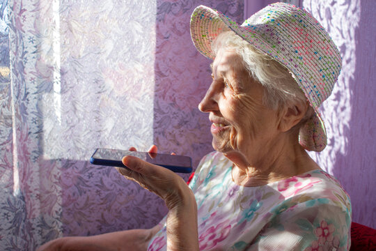 Charming elderly woman in rectangular black rimmed eyewear using voice recognition function on cell phone.