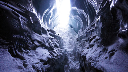 Light at the exit of the ice cave. A grotto in the mountains. The high ice walls are close by. The sun's rays do not get inside, they are reflected on the ice. There is white snow. October cave.