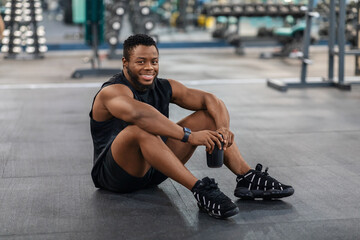 Smiling black guy in sportswear sitting on floor at gym
