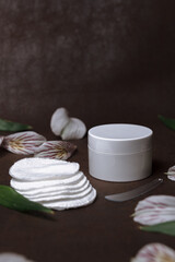 Organic facial cream and cotton pads placed with spatula and flower petals on table on brown background for natural skin care routine concept 