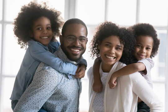 Candid lifestyle portrait of happy millennial African American married couple playing with two kids at home. Parents holding and piggy backing children, having fun smiling looking away. Family picture