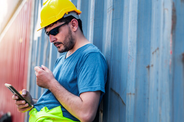 Young confident Caucasian man engineer using smartphone and wearing yellow safety helmet and check for control loading containers box from Cargo freight ship for import and export, transport.