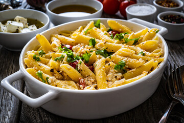 Roasted tomatoes with feta cheese and penne served in ceramic pan on wooden table