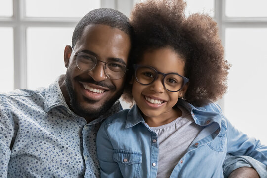 Portrait of happy African American dad and daughter girl both wearing glasses, hugging, relaxing, enjoying time together at home. Close up of father and kid sitting on sofa, looking at camera, smiling