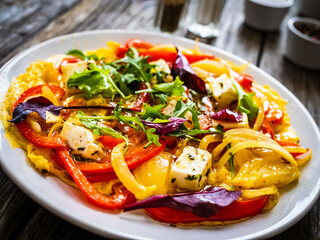 Delicious breakfast - scrambled eggs with onion, bell pepper, feta cheese and mix of young vegetable leaves on wooden table

