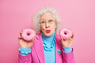 Lovely senior woman holds two glazed donuts keeps lips folded looks pleasantly at camera dressed in fashionable outfit with necklace and bracelet isolated over pink wall. Stylish granny with dessert