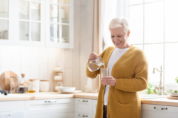 Happy senior woman pouring water into glass in kitchen - Powered by Adobe