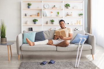 Millennial black guy with plastered leg sitting on sofa, reading book at home, pair of crutches standing nearby