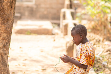 african kid using a smartphone. african child sitting alone outside using a phone and wearing...