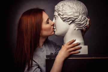 Red-haired young girl on a black background in a gray T-shirt kissing with a plaster bust