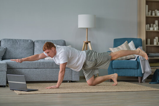 redhead man in white t-shirt works out in the living room. Man doing sports on a mat watching videos on a laptop in the living room with water and dumbbells on the floor