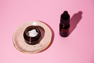 Open jar with cosmetic cream. Cosmetic dark amber glass bottle on pink background from above. Closeup, copyspace. Direct light. Beauty blogging, salon treatment concept, brand packaging mockup. 
